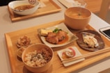 A wooden tray contains a variety of Japanese dishes, including grilled fish with sauce, a bowl of rice with sprinkles of seasoning, miso soup in a wooden bowl, a piece of grilled chicken with sauce and garnish on a ceramic plate, and small side dishes including pickled vegetables. Chopsticks rest on a small white ceramic holder, and a smartphone is partially visible on the table.