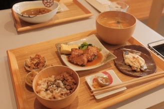 A wooden tray contains a variety of Japanese dishes, including grilled fish with sauce, a bowl of rice with sprinkles of seasoning, miso soup in a wooden bowl, a piece of grilled chicken with sauce and garnish on a ceramic plate, and small side dishes including pickled vegetables. Chopsticks rest on a small white ceramic holder, and a smartphone is partially visible on the table.