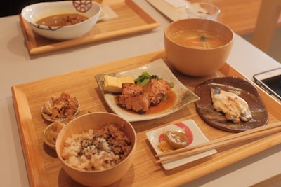 A wooden tray contains a variety of Japanese dishes, including grilled fish with sauce, a bowl of rice with sprinkles of seasoning, miso soup in a wooden bowl, a piece of grilled chicken with sauce and garnish on a ceramic plate, and small side dishes including pickled vegetables. Chopsticks rest on a small white ceramic holder, and a smartphone is partially visible on the table.