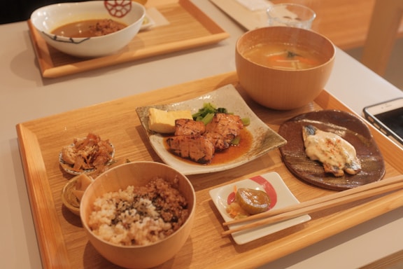 A wooden tray contains a variety of Japanese dishes, including grilled fish with sauce, a bowl of rice with sprinkles of seasoning, miso soup in a wooden bowl, a piece of grilled chicken with sauce and garnish on a ceramic plate, and small side dishes including pickled vegetables. Chopsticks rest on a small white ceramic holder, and a smartphone is partially visible on the table.