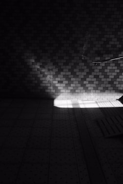 A moody, black and white photograph featuring a textured brick wall with a strong contrast of light and shadow creating geometric patterns. Sunlight streams in from the right, forming a bright spotlight on the floor which consists of a grid-like pattern with dotted tiles. A section of a railing or pipe is visible, leading off to the right.