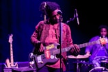 A musician is playing a bass guitar with stickers on it while standing on stage. The musician is dressed in a patterned shirt and jeans, and has headphones in their dreadlocks. In the background, another person is playing drums. The setting is dimly lit with a blue and purple hue.