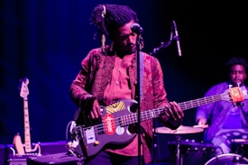 A musician is playing a bass guitar with stickers on it while standing on stage. The musician is dressed in a patterned shirt and jeans, and has headphones in their dreadlocks. In the background, another person is playing drums. The setting is dimly lit with a blue and purple hue.