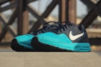 A pair of athletic running shoes with a turquoise front and a dark blue upper, featuring a prominent white logo on the side. The shoes are placed on a flat surface, with a blurred background showing a pattern of intersecting wooden beams.