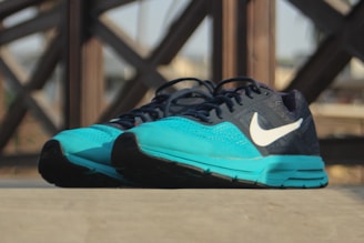 A pair of athletic running shoes with a turquoise front and a dark blue upper, featuring a prominent white logo on the side. The shoes are placed on a flat surface, with a blurred background showing a pattern of intersecting wooden beams.