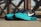 A pair of athletic running shoes with a turquoise front and a dark blue upper, featuring a prominent white logo on the side. The shoes are placed on a flat surface, with a blurred background showing a pattern of intersecting wooden beams.