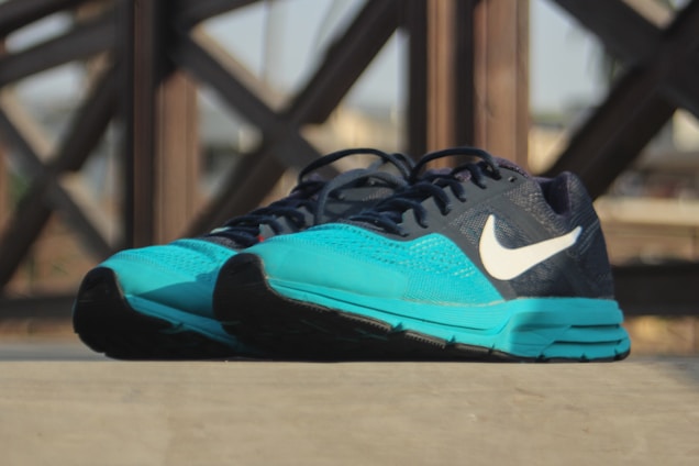 A pair of athletic running shoes with a turquoise front and a dark blue upper, featuring a prominent white logo on the side. The shoes are placed on a flat surface, with a blurred background showing a pattern of intersecting wooden beams.