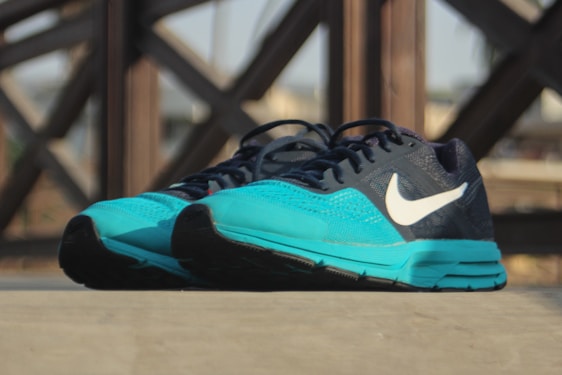A pair of athletic running shoes with a turquoise front and a dark blue upper, featuring a prominent white logo on the side. The shoes are placed on a flat surface, with a blurred background showing a pattern of intersecting wooden beams.