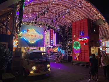A vibrant and colorful night scene featuring a retro-style van with 'Kombi Alley' signage illuminated by neon lights. Strings of lights decorate the arched roof above, creating a festival-like atmosphere. People are gathered around tables, suggesting a social or dining area. The setting is lively and energetic, with red shipping containers and additional neon signs adding to the eclectic decor.
