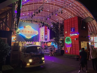 A vibrant and colorful night scene featuring a retro-style van with 'Kombi Alley' signage illuminated by neon lights. Strings of lights decorate the arched roof above, creating a festival-like atmosphere. People are gathered around tables, suggesting a social or dining area. The setting is lively and energetic, with red shipping containers and additional neon signs adding to the eclectic decor.
