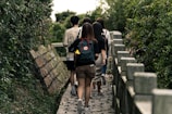 A group of people walking along a narrow stone pathway flanked by dense green foliage on both sides. The individuals are casually dressed, with one person carrying a backpack adorned with patches.