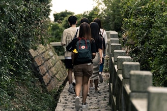 A group of people walking along a narrow stone pathway flanked by dense green foliage on both sides. The individuals are casually dressed, with one person carrying a backpack adorned with patches.