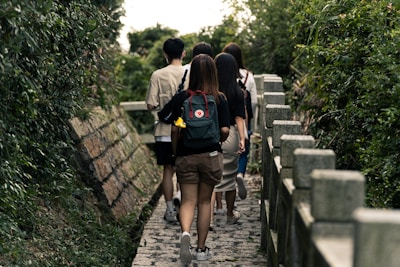 A group of people walking along a narrow stone pathway flanked by dense green foliage on both sides. The individuals are casually dressed, with one person carrying a backpack adorned with patches.
