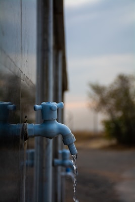 Several blue water taps are mounted on a wall with water flowing from one tap. The background features a blurred landscape with some trees and an overcast sky.
