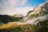 A scenic landscape features rugged mountains with rocky peaks and lush green valleys bathed in sunlight. A person with a backpack is hiking through the foreground, adding a sense of scale and adventure. The sky above is partly cloudy with patches of blue.