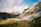 A scenic landscape features rugged mountains with rocky peaks and lush green valleys bathed in sunlight. A person with a backpack is hiking through the foreground, adding a sense of scale and adventure. The sky above is partly cloudy with patches of blue.