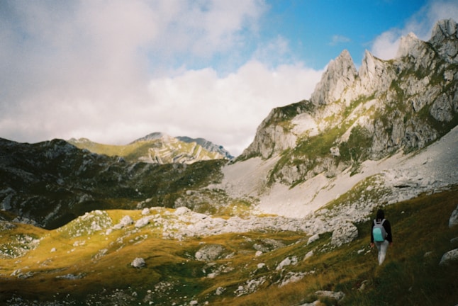 A scenic landscape features rugged mountains with rocky peaks and lush green valleys bathed in sunlight. A person with a backpack is hiking through the foreground, adding a sense of scale and adventure. The sky above is partly cloudy with patches of blue.