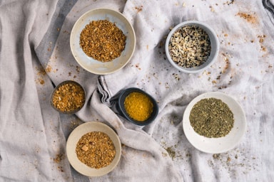 A collection of various spices displayed in small bowls on a textured cloth background. The spices range in colors and textures, including crushed seeds, dried herbs, and finely ground powders. The arrangement creates a natural and rustic culinary setting.
