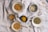 A collection of various spices displayed in small bowls on a textured cloth background. The spices range in colors and textures, including crushed seeds, dried herbs, and finely ground powders. The arrangement creates a natural and rustic culinary setting.