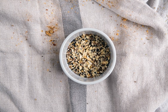 Close-up of a rustic bowl filled with talbeena plus dry fruit blend, surrounded by scattered nuts and grains on a wooden table.