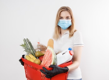 A person wearing a face mask holds a red shopping basket filled with groceries, including fruits, vegetables, and a large bottle, against a plain white background.
