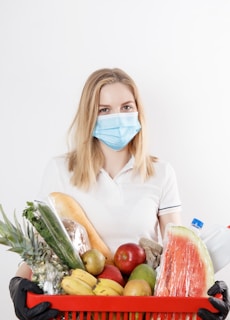 A person wearing a blue medical mask and a white polo shirt holds a red shopping basket full of groceries, including fresh fruits, vegetables, and a loaf of bread. Black gloves are worn by the person, indicating safety precautions.