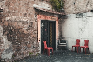 A rustic entrance to a pizzeria with a red sign above the door and flanking walls made of weathered brick. Three red chairs are positioned on a cobblestone surface to the right, accompanied by a distressed wooden bench. Lush greenery cascades from the roof, adding a touch of nature to the scene.