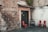 A rustic entrance to a pizzeria with a red sign above the door and flanking walls made of weathered brick. Three red chairs are positioned on a cobblestone surface to the right, accompanied by a distressed wooden bench. Lush greenery cascades from the roof, adding a touch of nature to the scene.