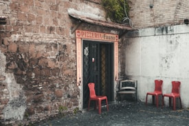 A rustic entrance to a pizzeria with a red sign above the door and flanking walls made of weathered brick. Three red chairs are positioned on a cobblestone surface to the right, accompanied by a distressed wooden bench. Lush greenery cascades from the roof, adding a touch of nature to the scene.