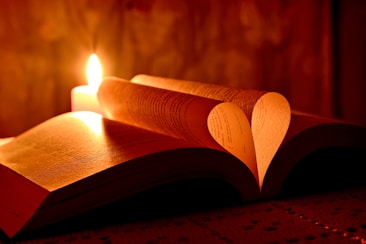 A dimly lit scene with an open book, where the pages are artfully folded to form a heart shape. A lit candle nearby casts a warm glow over the book, creating a cozy and intimate atmosphere.