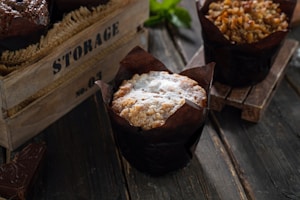 A rustic setting featuring a freshly baked muffin dusted with powdered sugar, presented in a dark paper liner. Surrounding it are additional muffins with visible nut toppings, set on a wooden surface that enhances the cozy, artisanal feel. A wooden box with the word 'STORAGE' and some stacked chocolate pieces complement the scene.