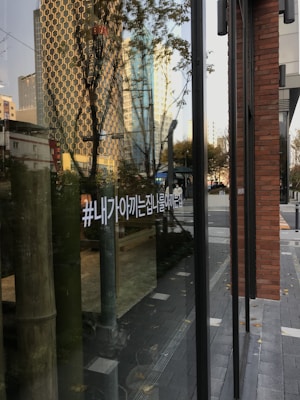 The image captures a reflection on a glass surface showcasing a cityscape with tall buildings in the background. Trees and urban elements, like a sidewalk and street, are visible. There is also a Korean hashtag written on the glass.