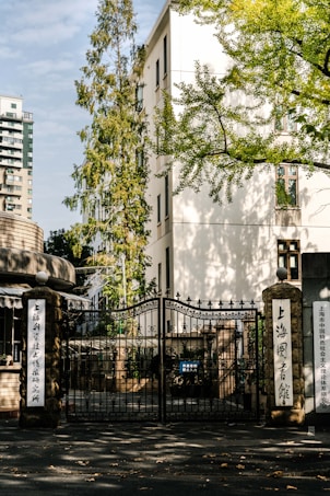 A wrought iron gate with intricate designs stands prominently in front of a light-colored building. The scene is flanked by tall stone pillars with inscriptions in Chinese. Trees with autumn foliage cast shadows across the pavement, and a modern apartment building is visible in the background.