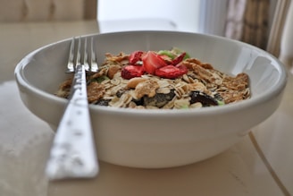 A white bowl filled with a variety of breakfast cereals including oats, flakes, and nuts, topped with slices of dried strawberries. A metal fork is placed in the bowl, resting along the edge.
