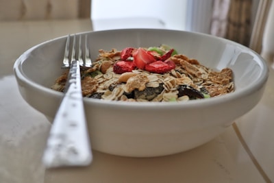 A white bowl filled with a variety of breakfast cereals including oats, flakes, and nuts, topped with slices of dried strawberries. A metal fork is placed in the bowl, resting along the edge.