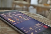 A smartphone is placed on a surface, displaying a live video or streaming interface with multiple user icons and text. The screen is slightly blurred, suggesting movement or focus on the background.