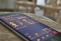 A smartphone is placed on a surface, displaying a live video or streaming interface with multiple user icons and text. The screen is slightly blurred, suggesting movement or focus on the background.