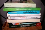 A stack of seven books is placed on a wooden surface with a pen lying beside it. The book covers display various colors and Arabic text. The titles span different genres, each with distinctive colors.