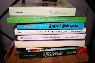 A stack of seven books is placed on a wooden surface with a pen lying beside it. The book covers display various colors and Arabic text. The titles span different genres, each with distinctive colors.
