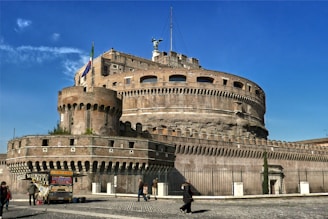 A massive cylindrical stone fortress with battlements and a large statue on top, standing under a clear blue sky. The structure is surrounded by a lower defensive wall and features a flag on a pole. Several people are walking along the stone pavement outside the fortress, and a couple of small vendor stalls are set up nearby.