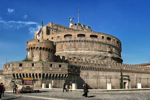 A massive cylindrical stone fortress with battlements and a large statue on top, standing under a clear blue sky. The structure is surrounded by a lower defensive wall and features a flag on a pole. Several people are walking along the stone pavement outside the fortress, and a couple of small vendor stalls are set up nearby.