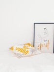 A minimalist interior featuring a pillow with yellow and gray leaf patterns and a framed artwork depicting a simple line drawing of a face with the words 'free spirit'. The setting is bright and clean with a white background.