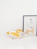 A minimalist interior featuring a pillow with yellow and gray leaf patterns and a framed artwork depicting a simple line drawing of a face with the words 'free spirit'. The setting is bright and clean with a white background.
