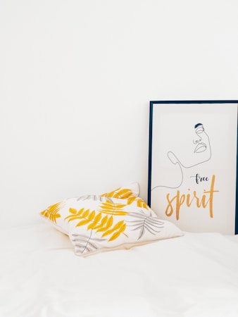 A minimalist interior featuring a pillow with yellow and gray leaf patterns and a framed artwork depicting a simple line drawing of a face with the words 'free spirit'. The setting is bright and clean with a white background.