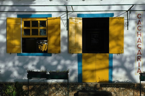 A quaint storefront with bright yellow shutters and door, framed by vibrant blue trim. The word 'Cachacaria' is vertically displayed on the right side. The facade is adorned with shadows of nearby foliage, enhancing the rustic charm.