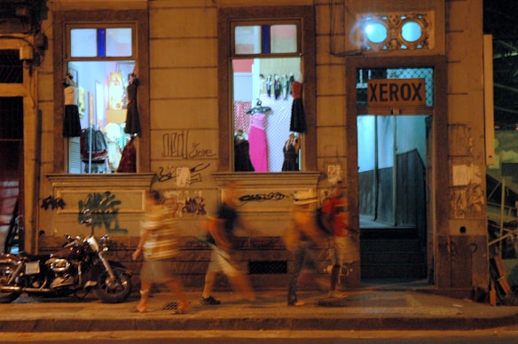 An urban nighttime scene features a storefront with brightly lit windows displaying mannequins wearing dresses. Below the windows, a motorcycle is parked on the street, and graffiti adorns the building's façade. A group of people walks by, captured with motion blur, indicating movement.