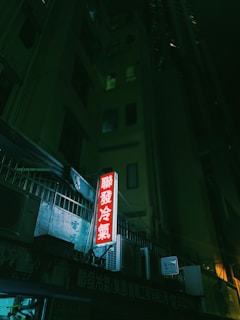 Neon signage in a dark urban alley with a prominent red glow against the building's grim facade. Several air conditioning units are mounted on the exterior, adding to the urban setting.