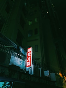 Neon signage in a dark urban alley with a prominent red glow against the building's grim facade. Several air conditioning units are mounted on the exterior, adding to the urban setting.