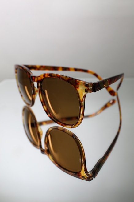 A pair of tortoiseshell sunglasses with brown tinted lenses is positioned against a reflective surface, creating a clear mirror image. The frame has a glossy finish, adding a touch of sophistication.