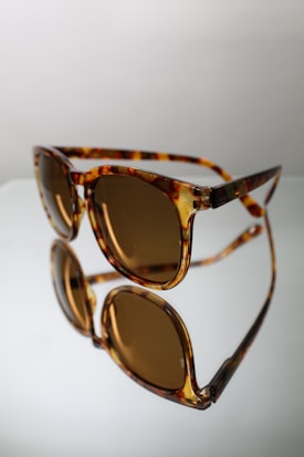 A pair of tortoiseshell sunglasses with brown tinted lenses is positioned against a reflective surface, creating a clear mirror image. The frame has a glossy finish, adding a touch of sophistication.